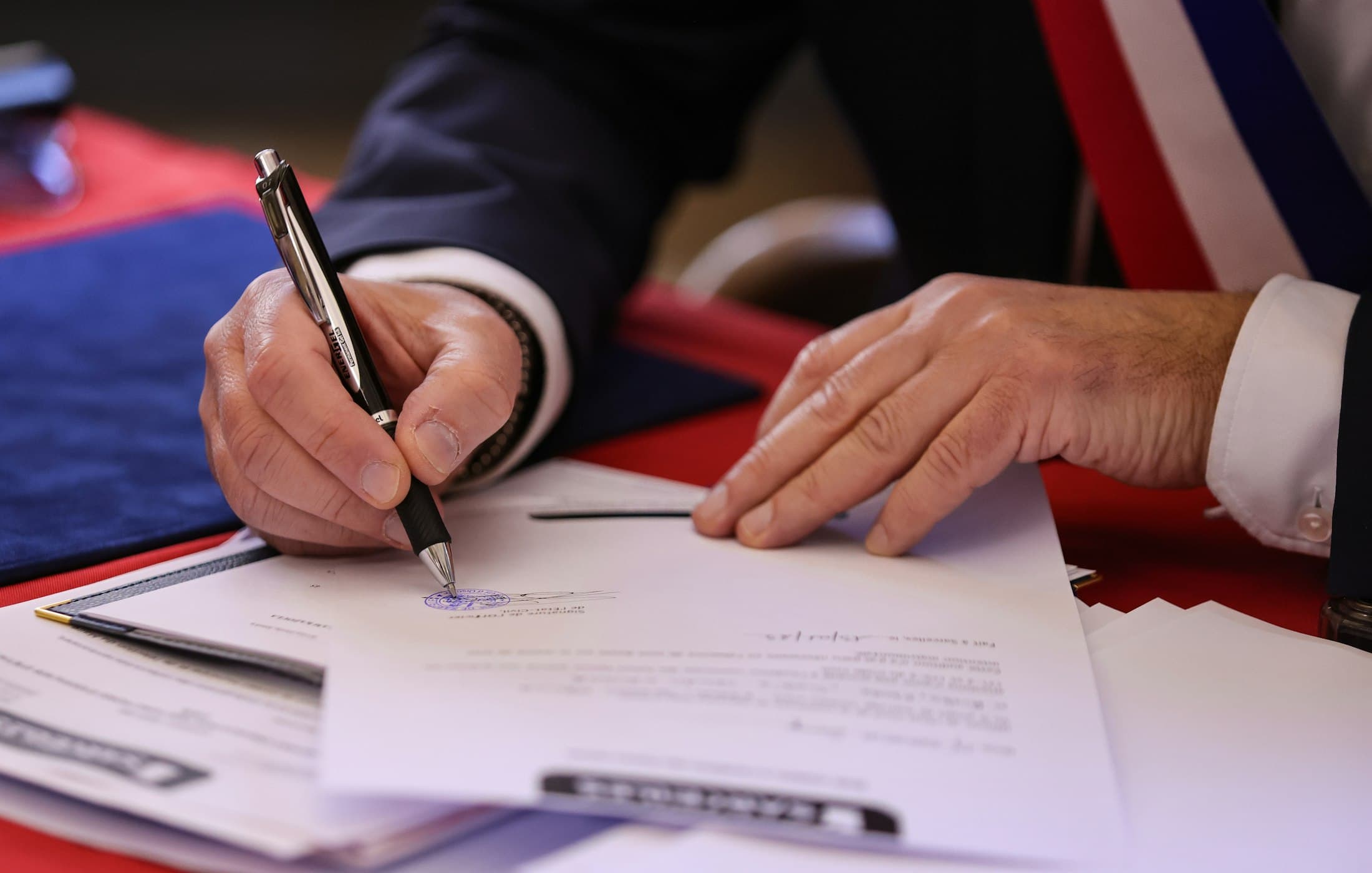 Main en costume signant un document relié avec tampon prêt et annexes alignées sur le bureau